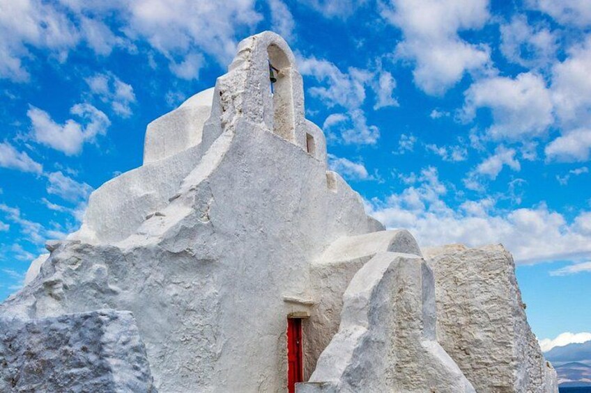 En hvid bygning af sten med en rød dør og et vindue, mod en blå himmel med skyer. 2