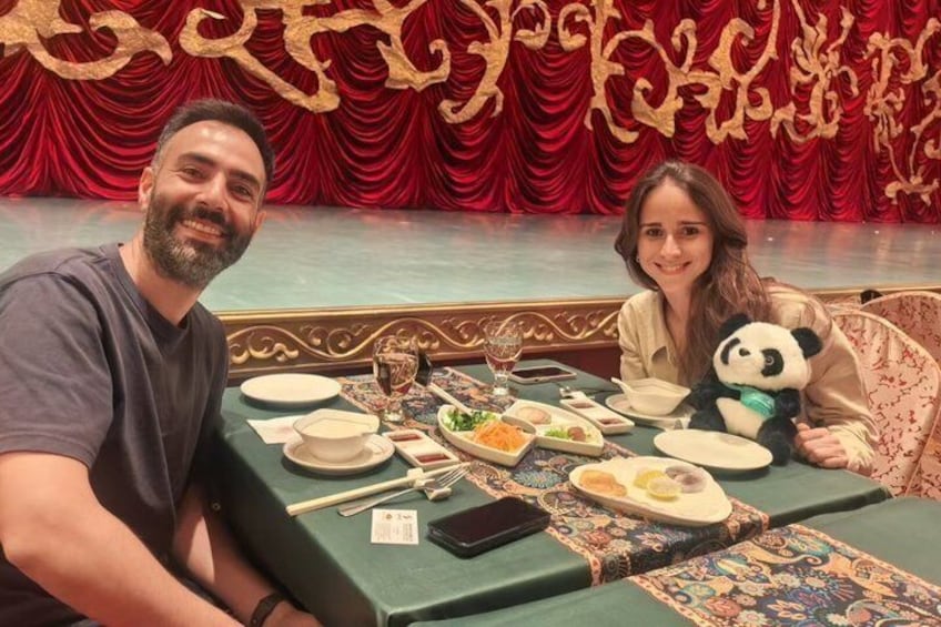 Twee personen zitten aan een tafel met diverse gerechten, een pluche panda en een rode gordijn achtergrond. 2