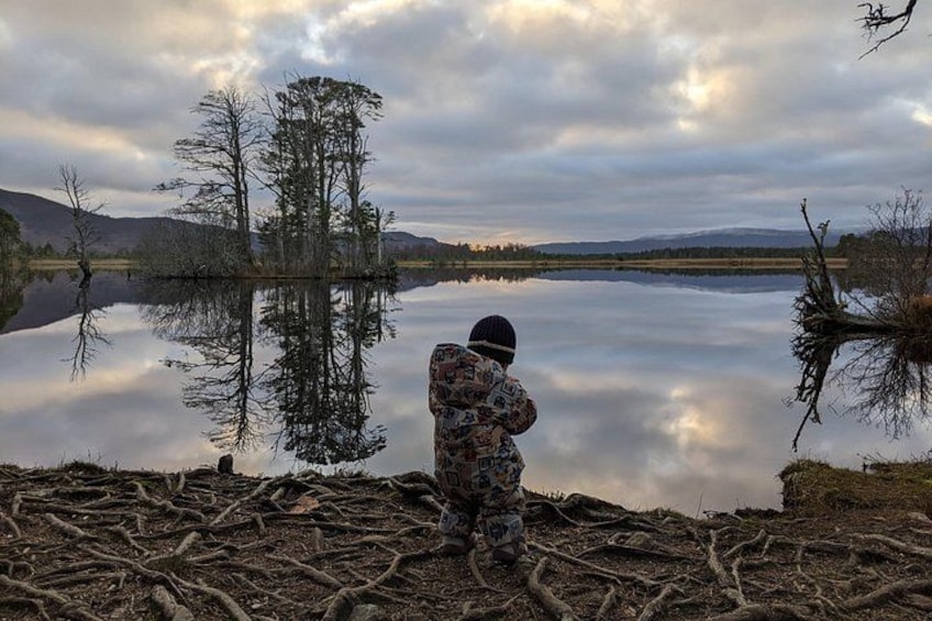 Eine Person in Tarnkleidung steht an einem stillen See, dessen Oberfläche Bäume und Berge widerspiegelt. 2
