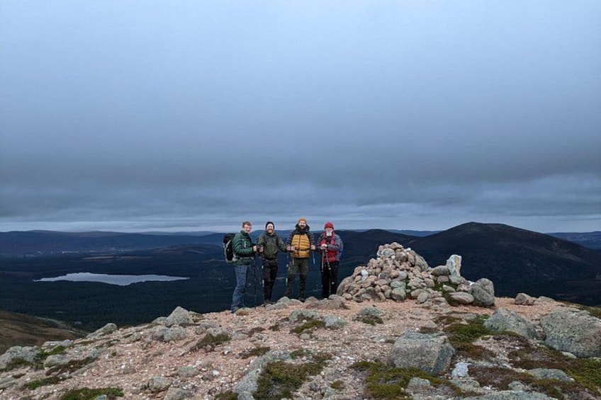 Eine Gruppe Wanderer auf einem felsigen Berggipfel, im Hintergrund ein See und bewölkter Himmel. 4