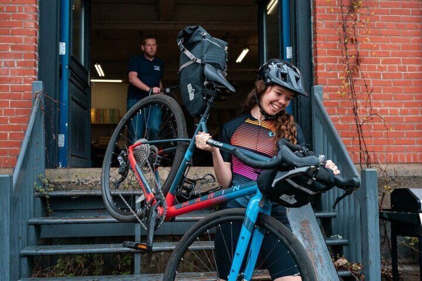 A person on a blue bicycle with a black bag on the back, standing on steps in front of a red brick building. 3