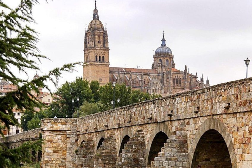 A stone bridge with arches, a historic building with a bell tower and dome, and trees in the foreground. 1