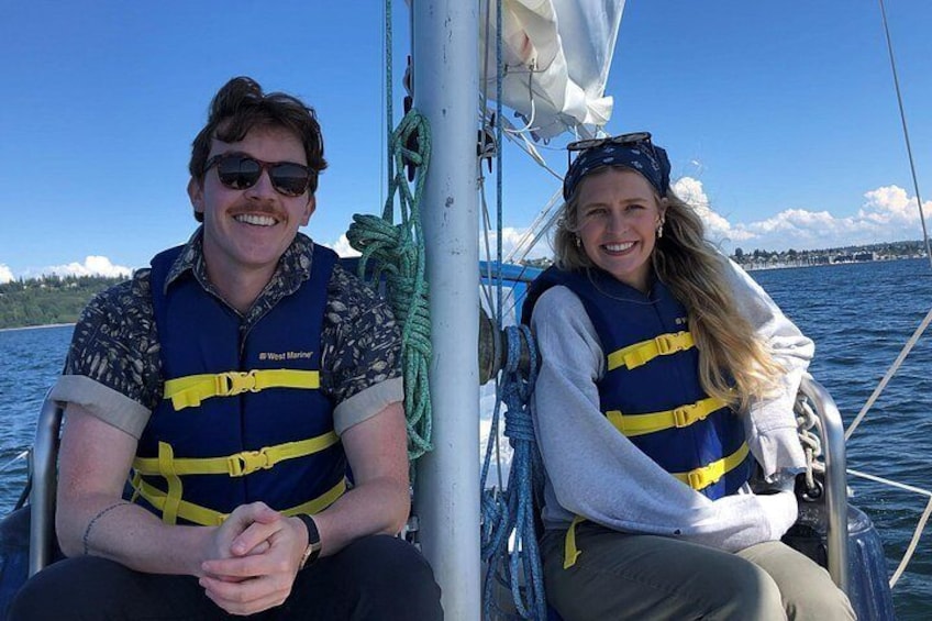 Twee mensen op een zeilboot, allen met reddingsvesten, met een wateroppervlak en een verre kustlijn op de achtergrond. 3