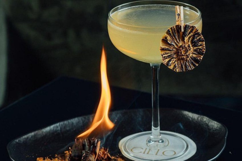A cocktail glass with a garnish on a plate labeled 'CILIO' next to a lit fire. 4