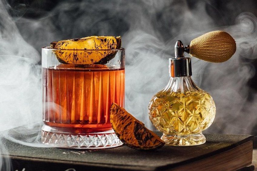 A glass of amber liquid with a peel, a bottle with a textured cap, and a book on a table. 3