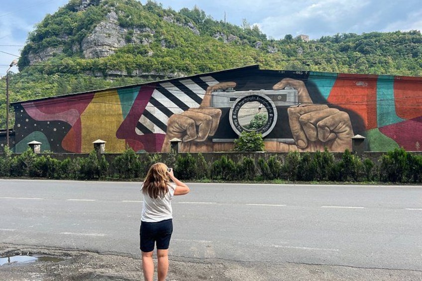 Un murale su un edificio raffigura una macchina fotografica, mani e una donna che scatta una foto, con una strada sullo sfondo. 1
