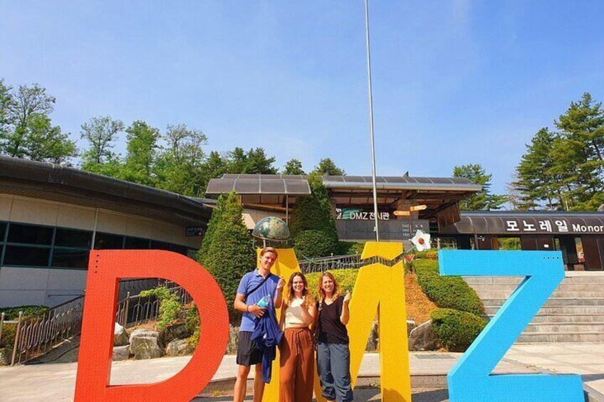 A large outdoor sign with the letters 'DMZ' in bold colors, a building with a sign reading 'Monorail', and a flagpole with a flag. 1