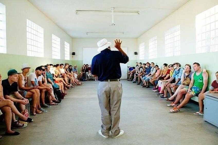 A person addressing a seated audience in a room with large windows. 2