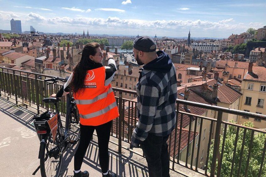 Een persoon in een oranje vest staat naast een fiets op een balkon met uitzicht op een stadsgezicht. 3