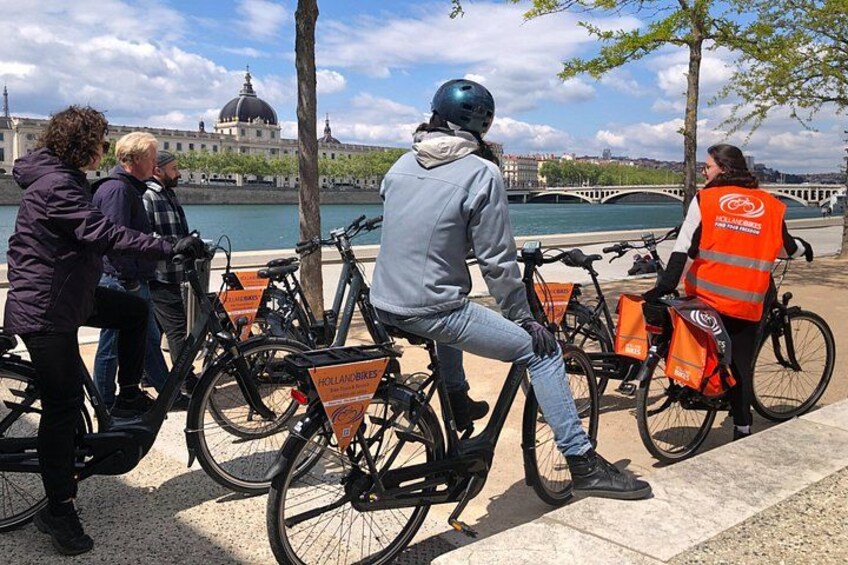 Een groep mensen op fietsen nabij een rivier, met een brug op de achtergrond. 2