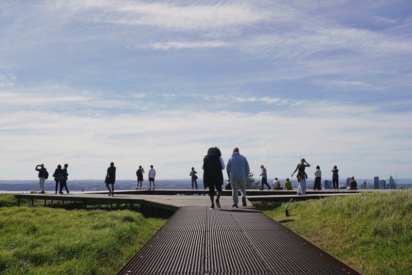 En træpromenade med gående mennesker, grønne områder og en bysilhuet i det fjerne. 4