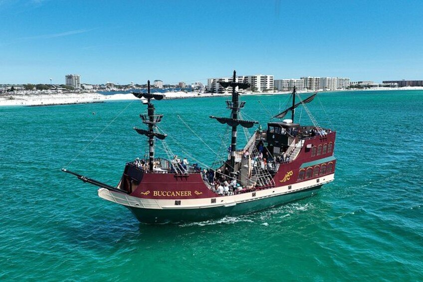 Un gran barco de vela histórico llamado 'Buccaneer' en el agua, con una playa y edificios al fondo. 2