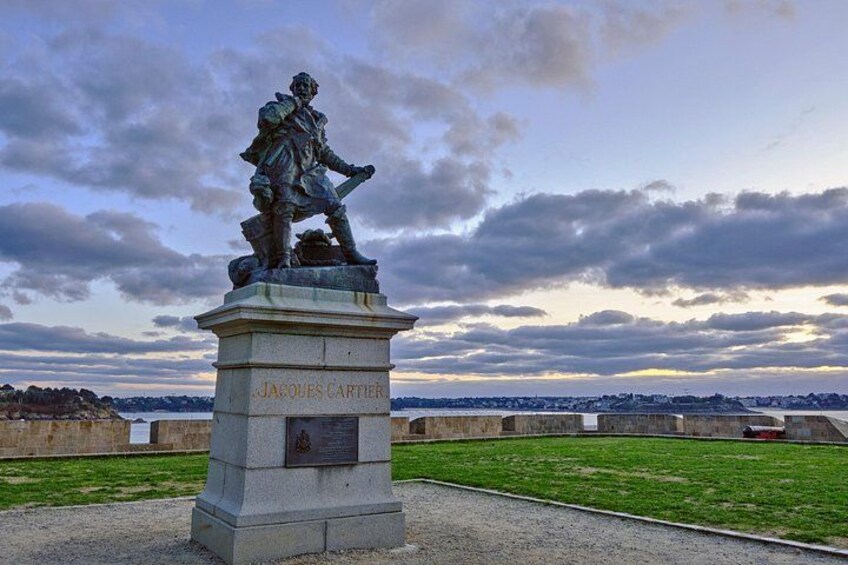 Une statue de Jacques Cartier, posée sur un piédestal et surmontée d’une plaque commémorative, se détache sur un arrière-plan composé d’un ciel nuageux et d’une vue lointaine de la ville. 2