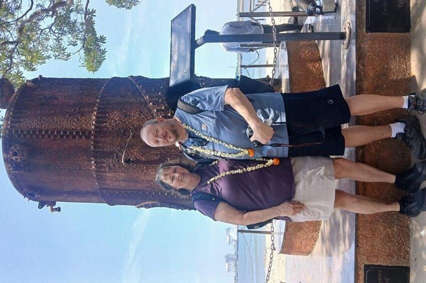 Two people standing in front of a large, rusted metal structure with a chain and a plaque. 2