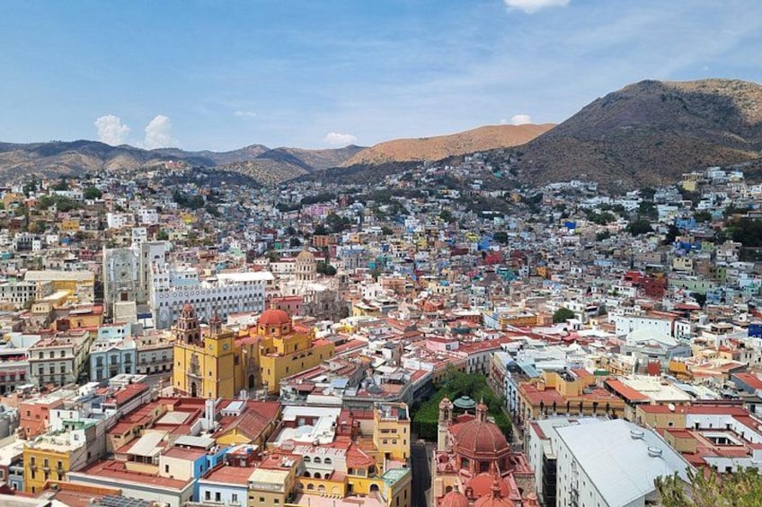 Un paisaje urbano denso con edificios coloridos, una iglesia amarilla destacada y una montaña al fondo. 1