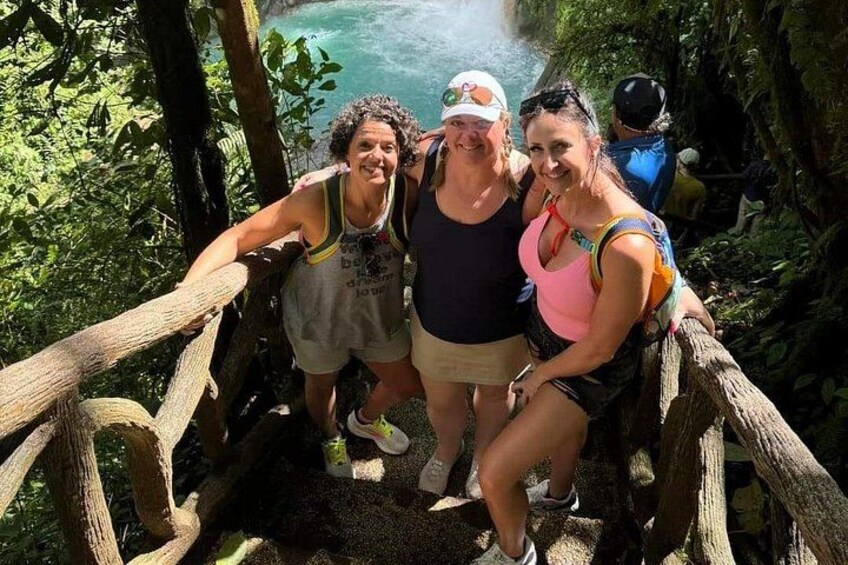 Un groupe de personnes se trouve sur un pont en bois, dans une zone boisée, avec une étendue d'eau en arrière-plan. 1