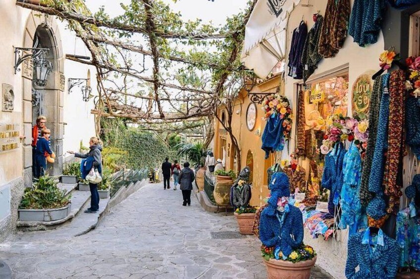 Une ruelle étroite pavée, bordée de boutiques, dont certaines exposent à l’extérieur des étoffes colorées et des fleurs. 3