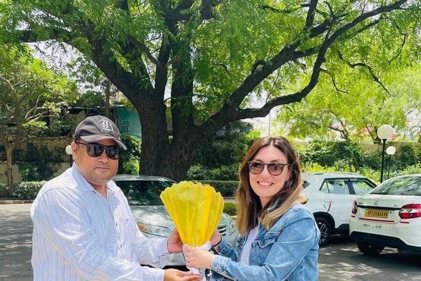 Dos personas sostienen flores amarillas en un estacionamiento, con coches y un árbol grande al fondo. 4