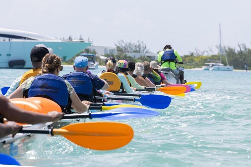 A group of people rowing in a boat on a body of water with a boat and a person in a kayak in the background. 2