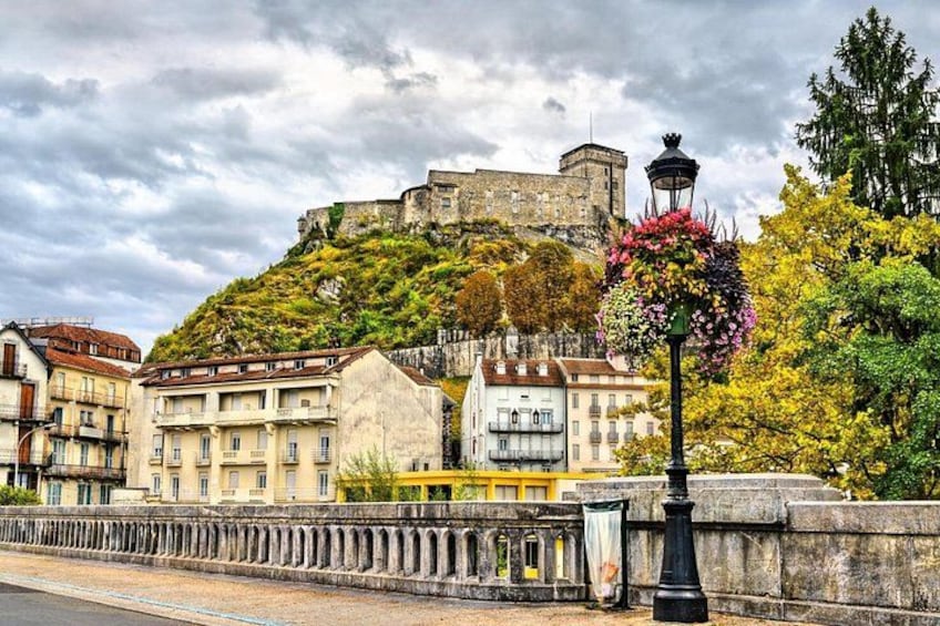 A historic cityscape with a prominent castle on a hill, a bridge with a railing, and a street lamp with flowers. 1