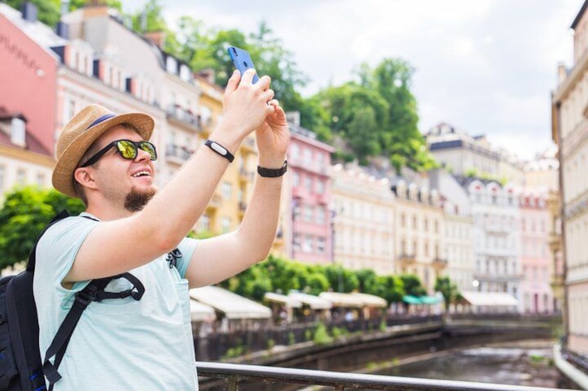 A person taking a selfie with a smartphone, wearing a straw hat and a light blue shirt, with a backpack, in a city with colorful buildings and a canal in the background. 3