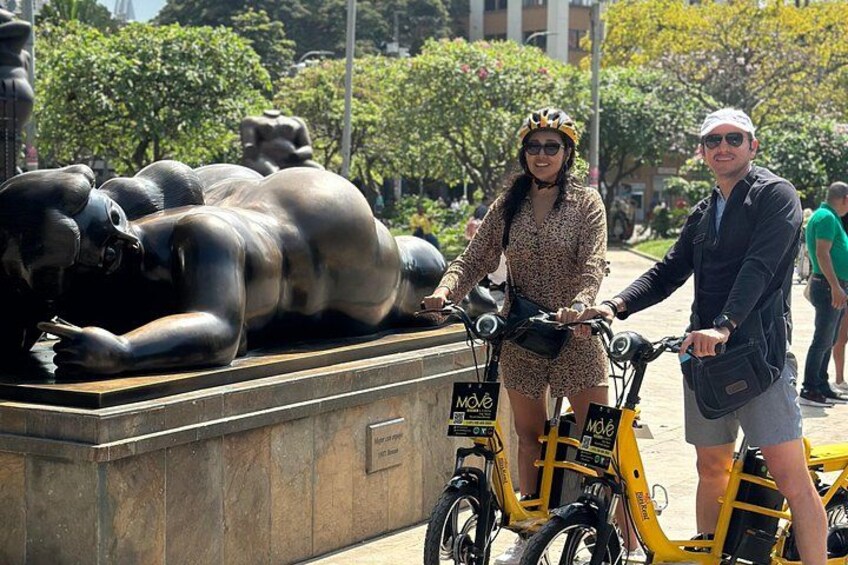 A sculpture of reclining figures, a person on a yellow bicycle, and another person standing beside it. 1