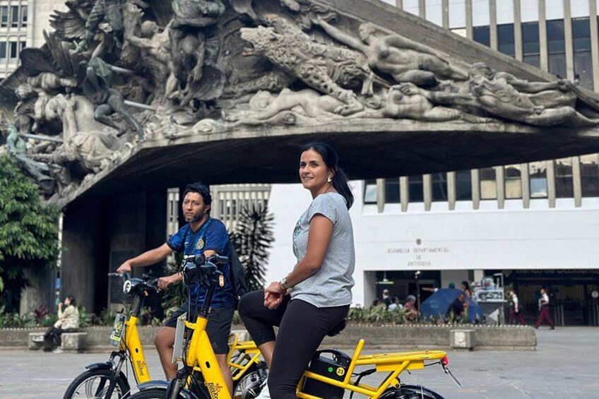 Two people on a yellow electric bike, a sculpture, and a building with signage. 3