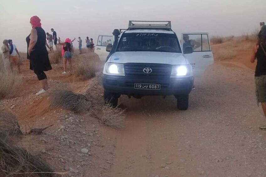 A white SUV on a dirt road with people standing on the side. 2