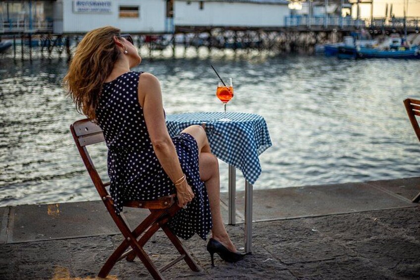A woman sitting on a chair by a waterfront, wearing a polka dot dress and high heels, with a table and a drink in front of her. 2