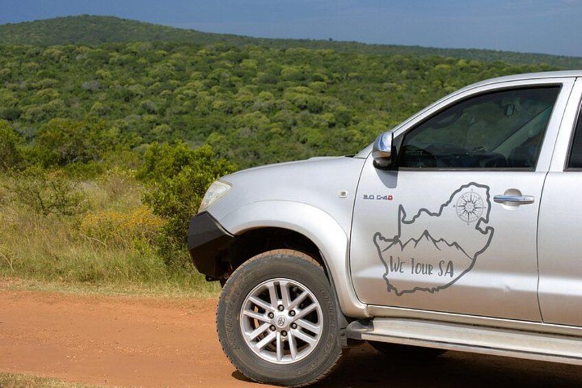 A silver SUV with a map decal on the side door, parked on a dirt road with green vegetation and hills in the background. 4