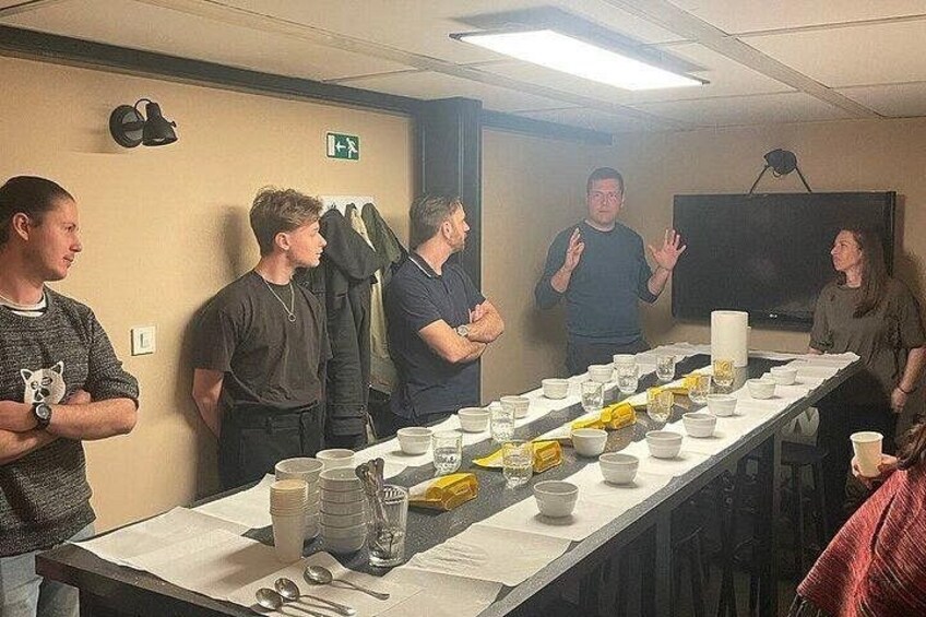 Un grupo de personas de pie alrededor de una mesa larga con tazones y tazas, en una habitación con una pizarra y un reloj en la pared. 3