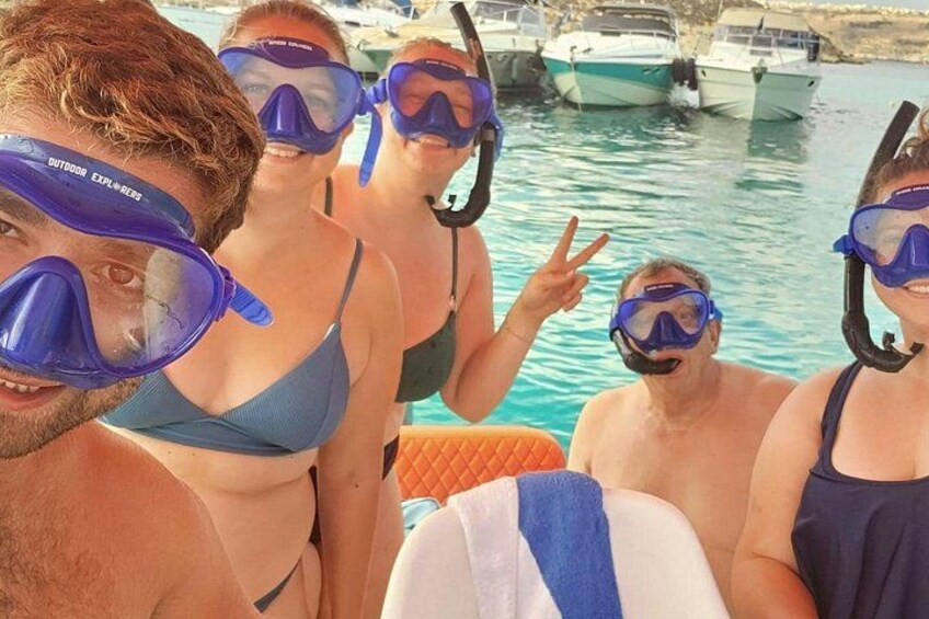 Eine Gruppe von Menschen schnorchelt mit blauen Masken und Flossen, im Hintergrund Boote und eine felsige Küste. 3