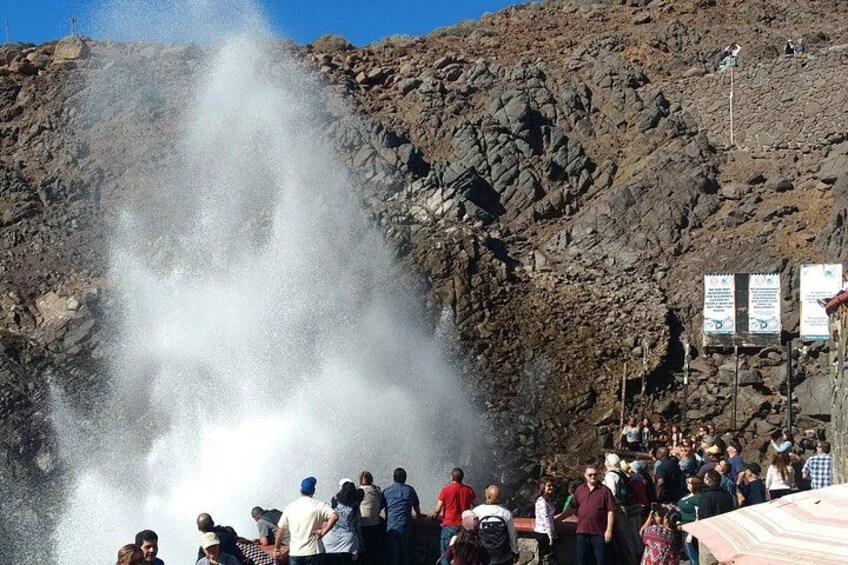 Une foule observe l’éruption d’un geyser, avec un paysage rocheux et des panneaux indicateurs en arrière-plan. 3