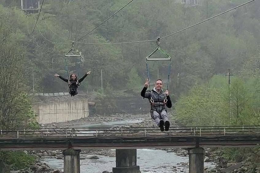 Twee mensen op een zipline boven een rivier, met een brug en groen in de achtergrond. 5