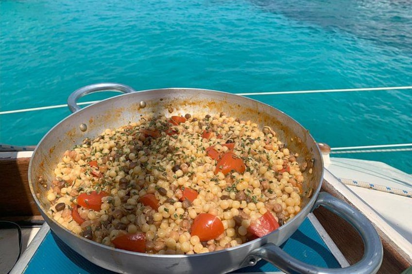 Een grote metalen pan gevuld met een gerecht met kikkererwten, tomaten en kruiden, op een boot met een blauwe reling. 1