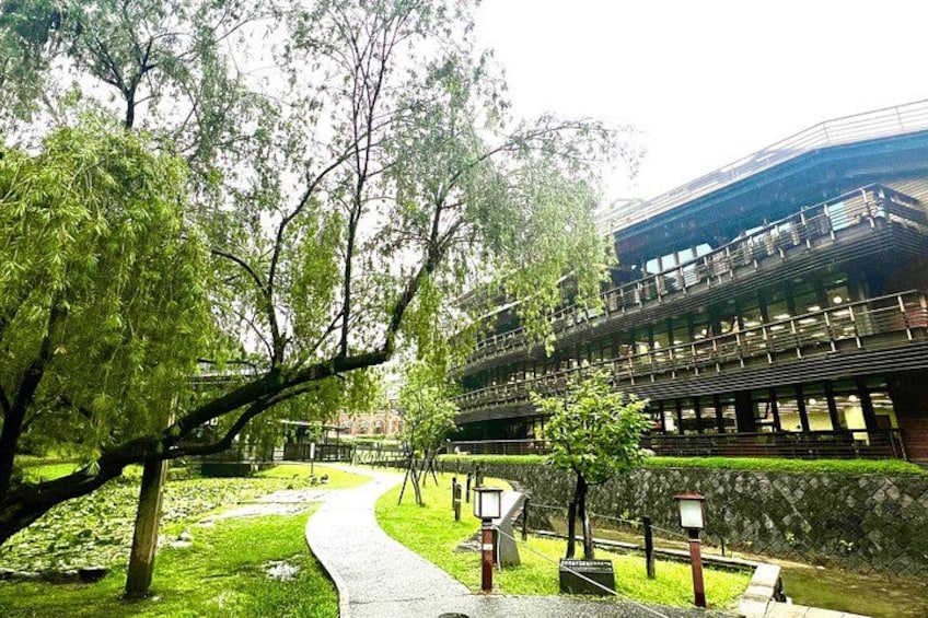 A pathway leading to a modern building with a glass facade, surrounded by trees and a small pond. 5