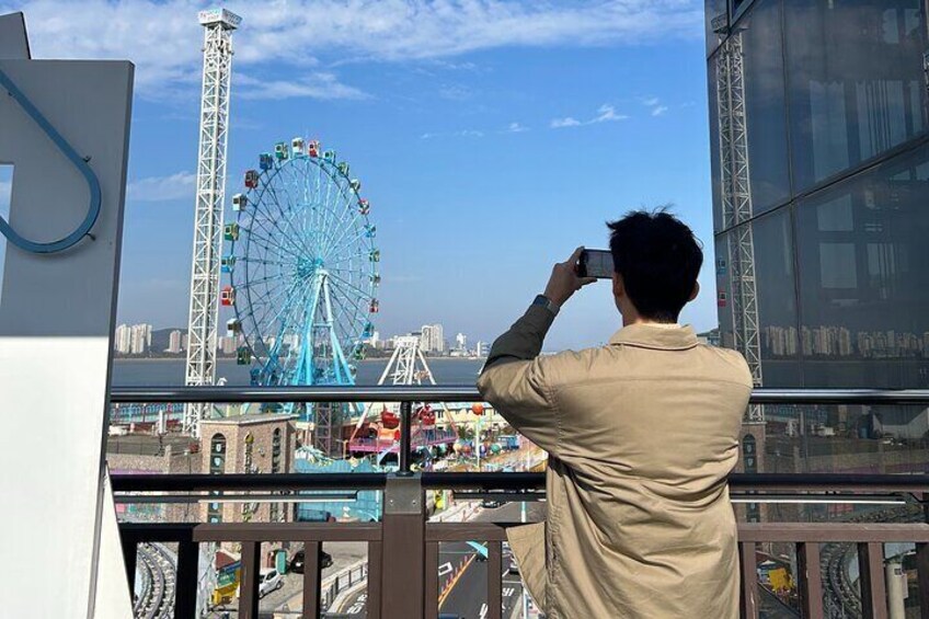 A person taking a photo of a Ferris wheel from a balcony overlooking a cityscape. 1