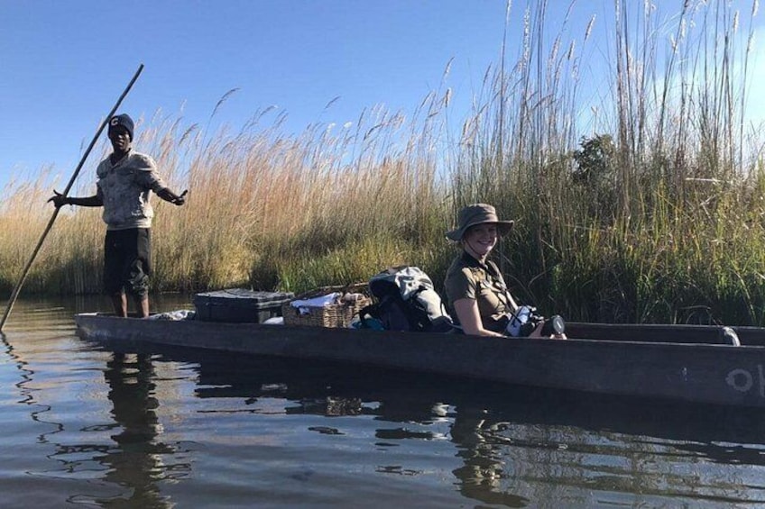 Two people in a narrow waterway, one standing and one seated, with a canoe and gear. 3