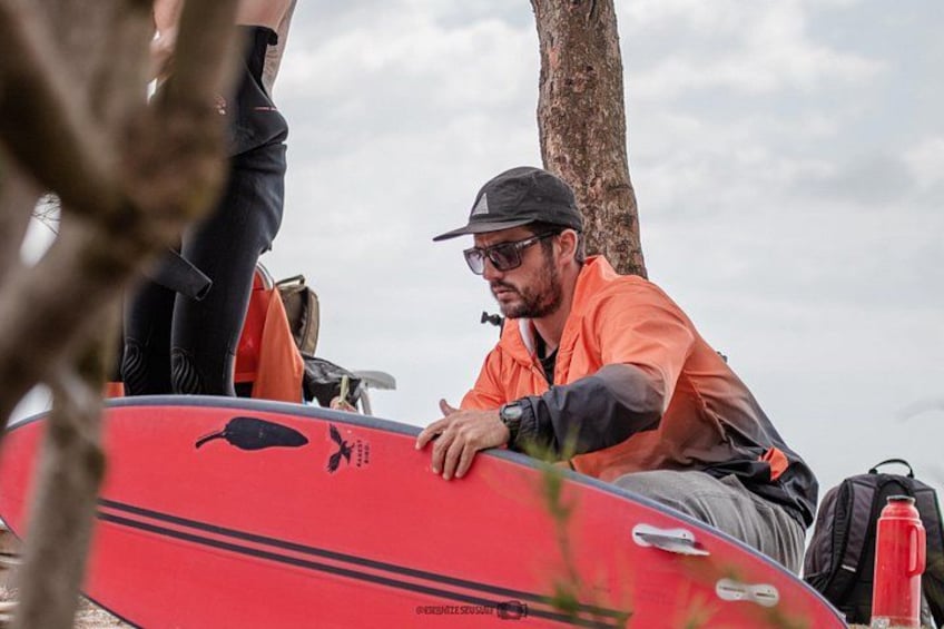 A person sitting on a red surfboard with a black whale design, wearing an orange shirt and a cap, with a tree and a cloudy sky in the background. 2