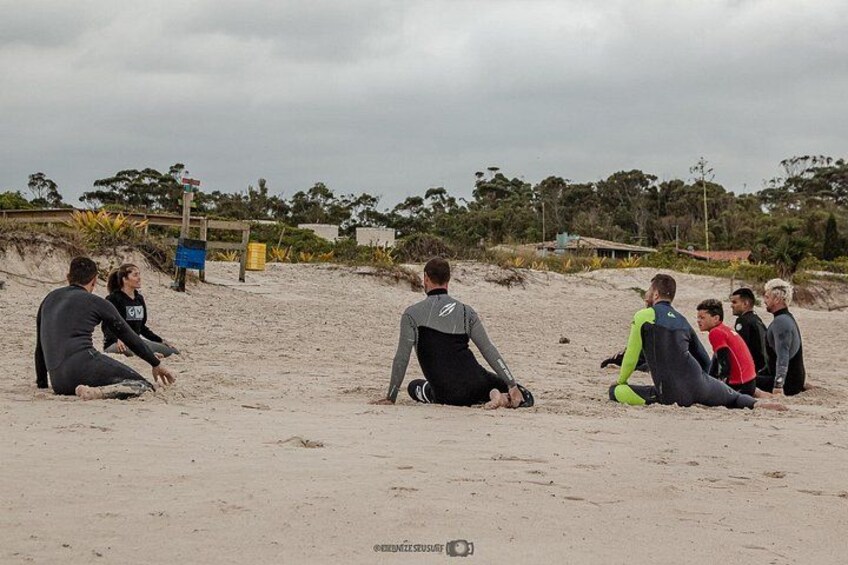 Eine Gruppe von Menschen sitzt auf einem Sandstrand, manche tragen Neoprenanzüge. 2