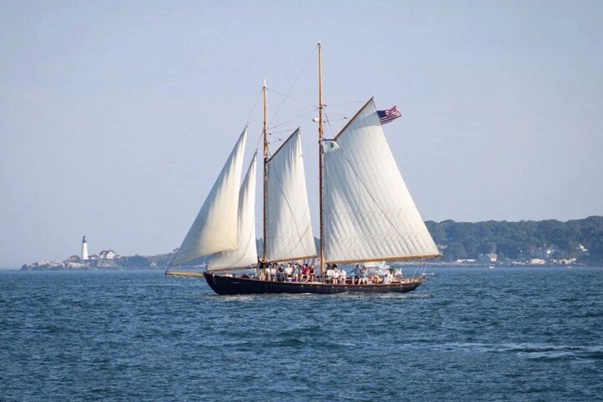 A sailboat with multiple sails on a body of water, a lighthouse in the background, and a coastline with buildings. 3
