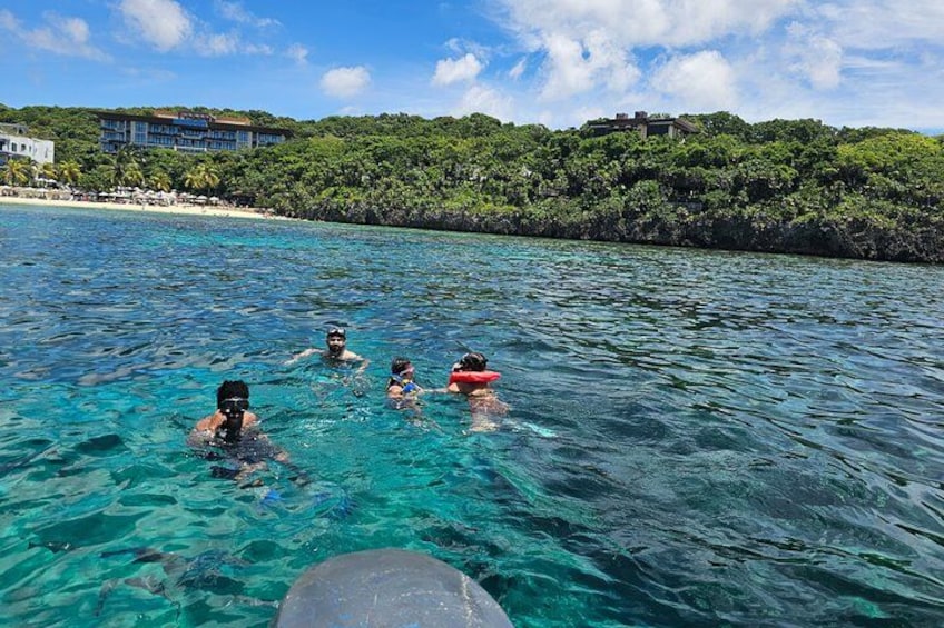 Personas nadando en aguas turquesas cristalinas, con una playa y exuberante vegetación al fondo. 2