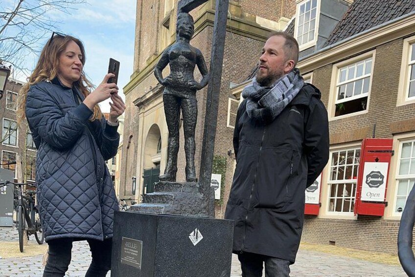 Two people standing in front of a statue, with a red mailbox and a building in the background. 1