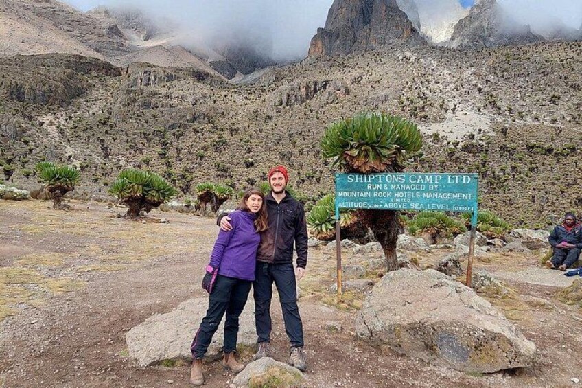 A couple posing in front of a sign for Shiptons Camp Ltd, with a mountainous landscape in the background. 4