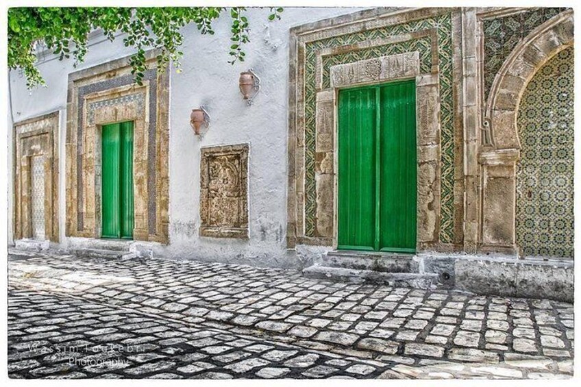 A cobblestone street with white buildings, green doors, and decorative stonework. 3