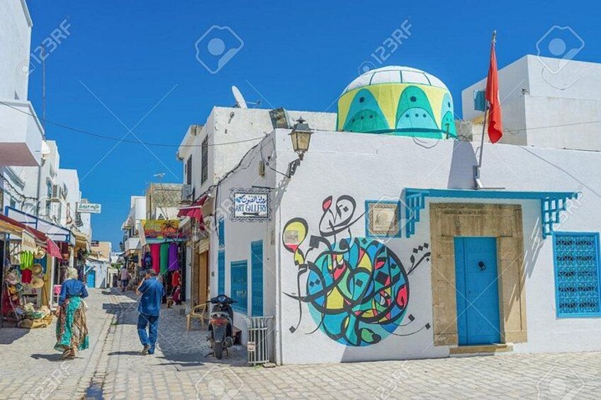 A narrow street with white buildings, colorful murals, and a blue-domed structure. 1