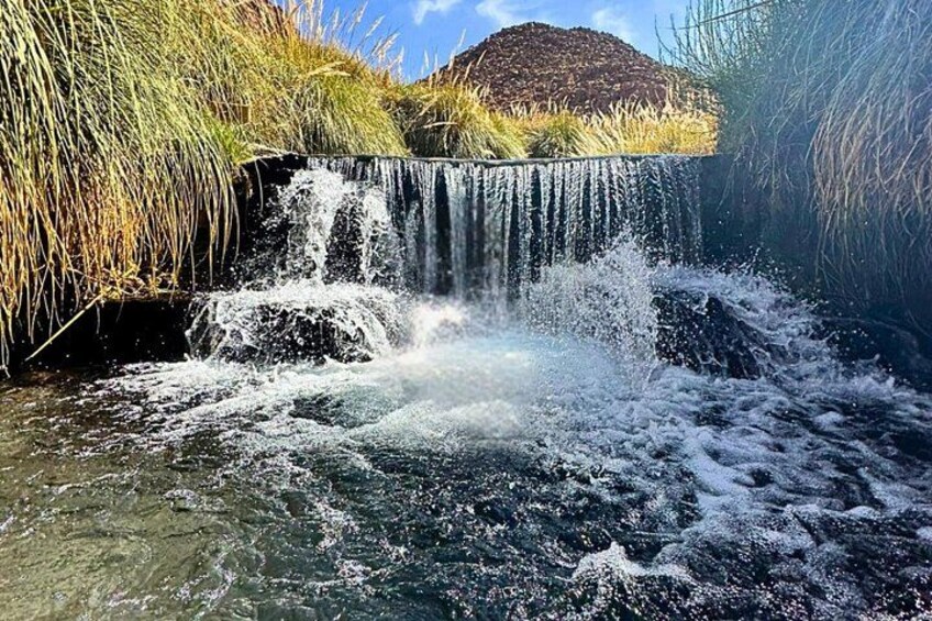 Een waterval stort neer over rotsen, omgeven door hoog gras en een heuvel op de achtergrond. 5