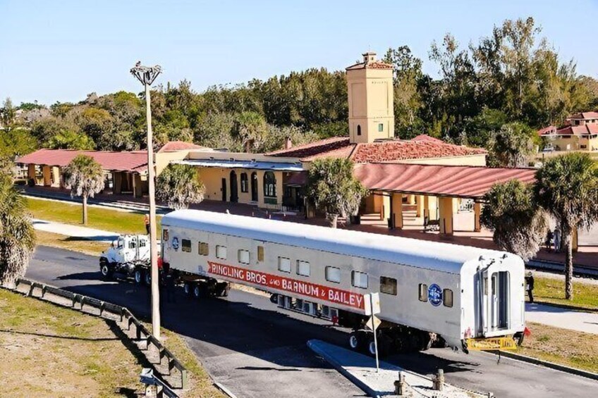 Een oude trein met 'RINGLING BROS. BAR NUM & BOILERY' op de zijkant, geparkeerd op een straat met gebouwen en bomen op de achtergrond. 1