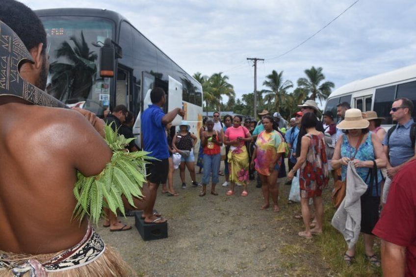 A group of people standing in a line, some holding items, with a bus and palm trees in the background. 3