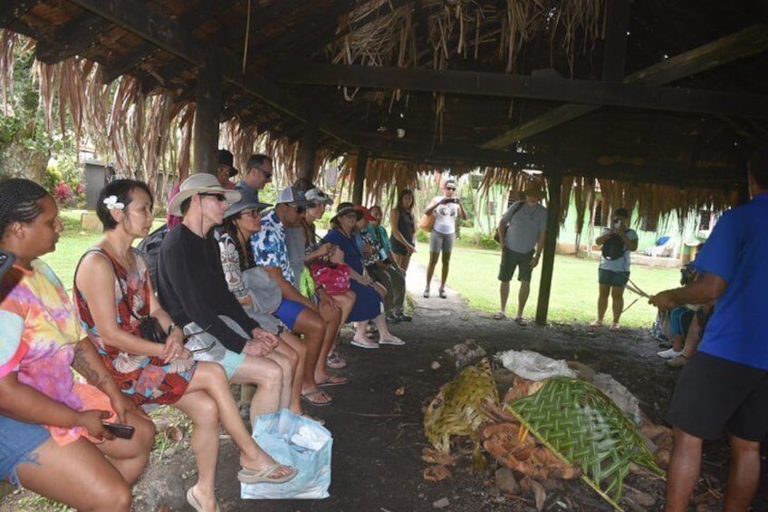 People seated under a thatched roof structure, observing a person preparing food outdoors. 5